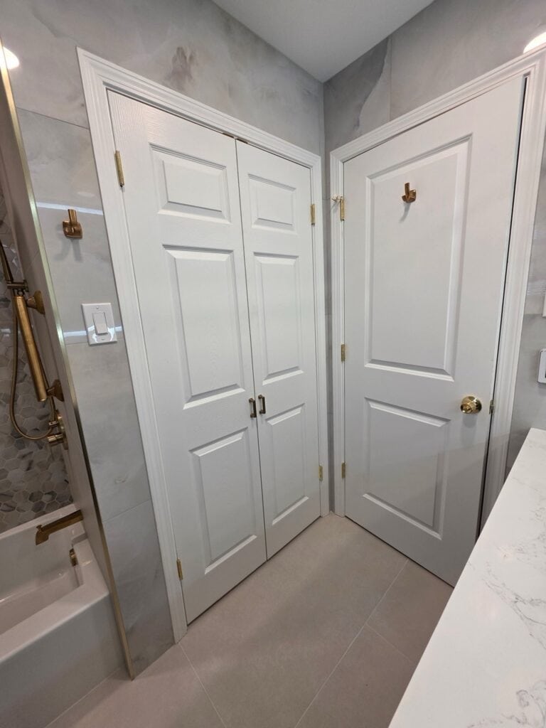 Modern bathroom with white double doors and gold hooks, marble walls, and a bathtub.
